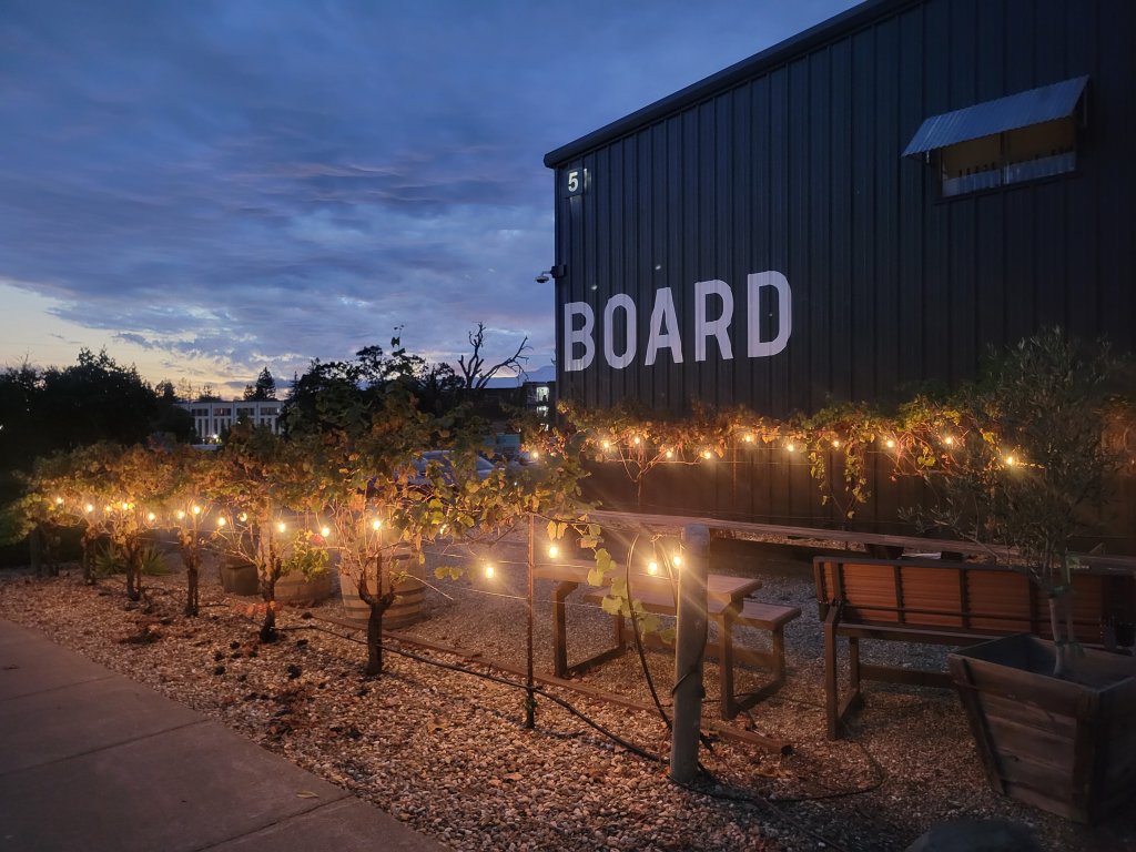 Evening view of a black building with 'BOARD' sign, surrounded by lit plants and benches in Healdsburg, California.