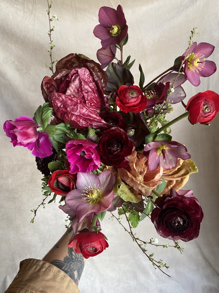 A vibrant bouquet of deep red, purple, and pink flowers, including roses, ranunculus, and hellebores, held by a hand with a tattooed sleeve against a neutral background in Healdsburg, California.