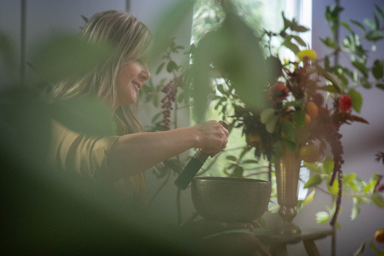 A woman is holding a bowl and a mallet, surrounded by lush greenery and vibrant flowers near a window in Healdsburg, California.
