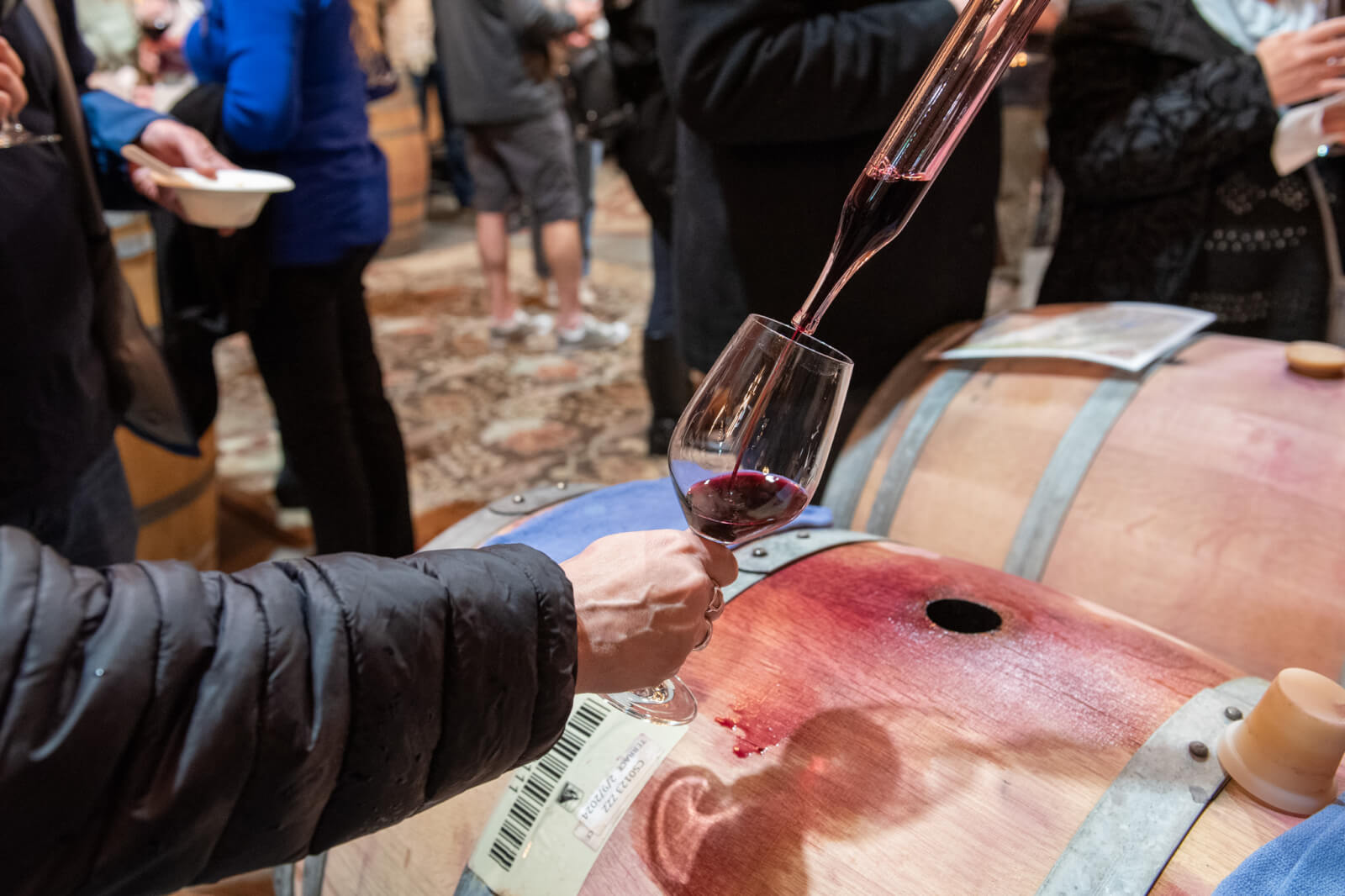 A person pours red wine from a decanter into a glass held over a wooden barrel at a wine tasting event in Healdsburg, California.