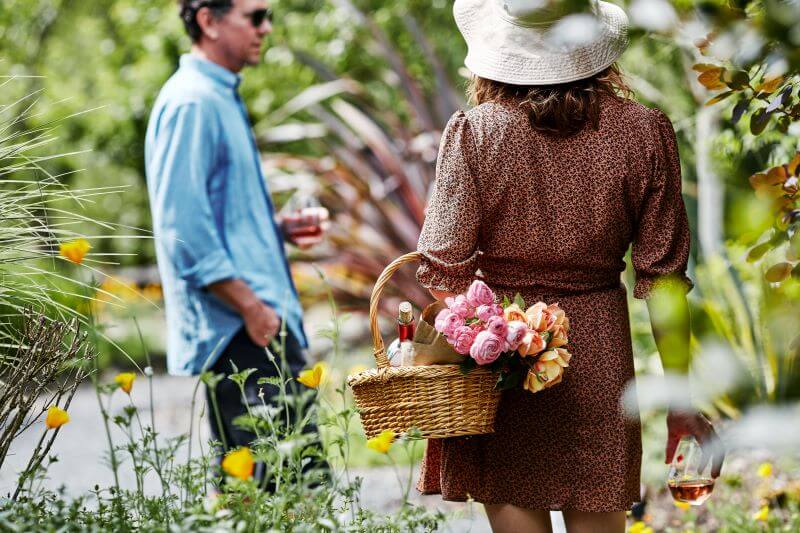 A woman in a floral dress and wide-brimmed hat carries a basket of pink roses through a sunny garden, while a man in a blue shirt and sunglasses walks behind her, holding a glass of wine in Healdsburg, California.