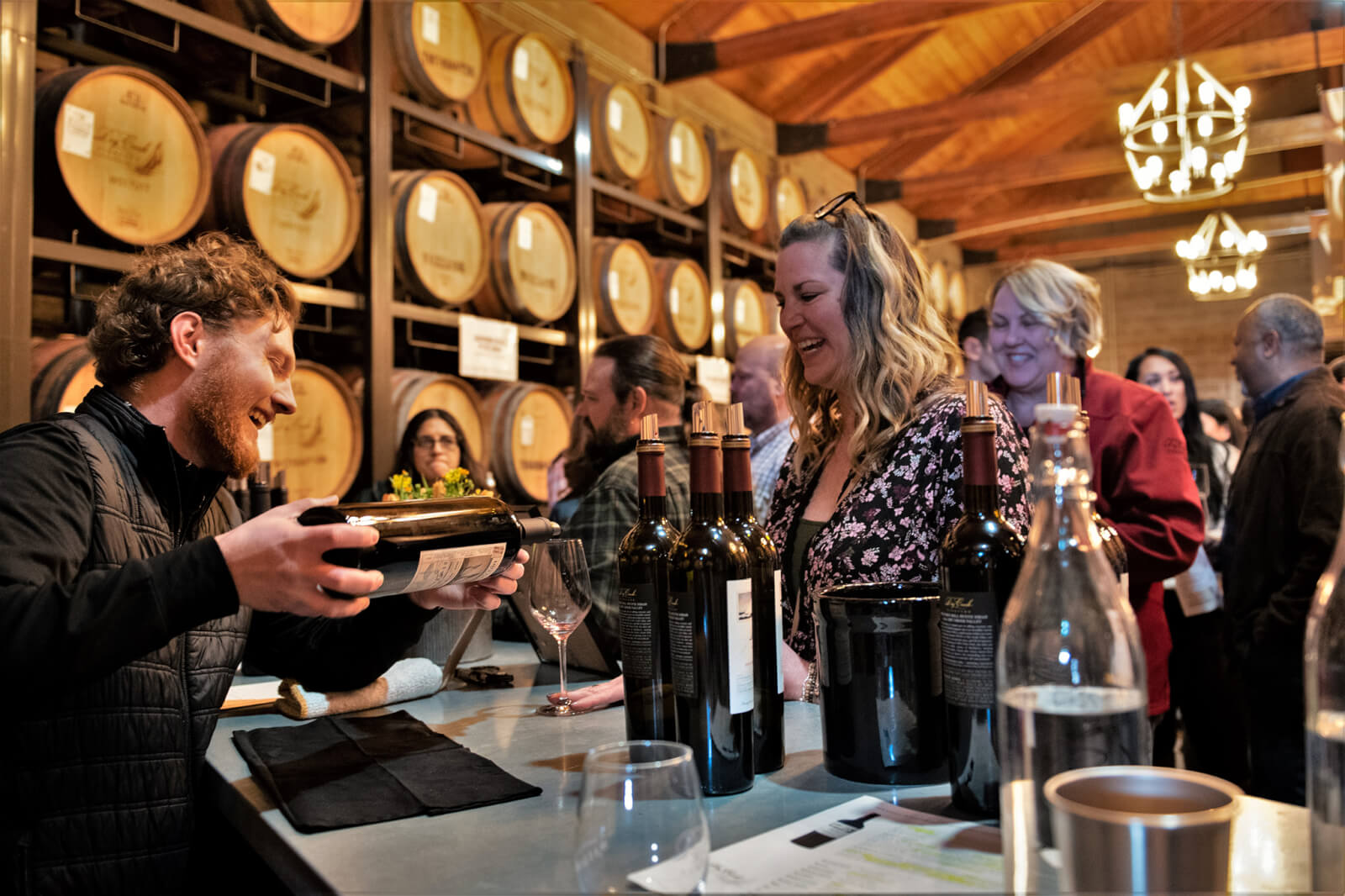 A lively wine tasting event in a cozy cellar with wooden barrels and chandeliers, where guests sample various wines and enjoy each other's company in Healdsburg, California.