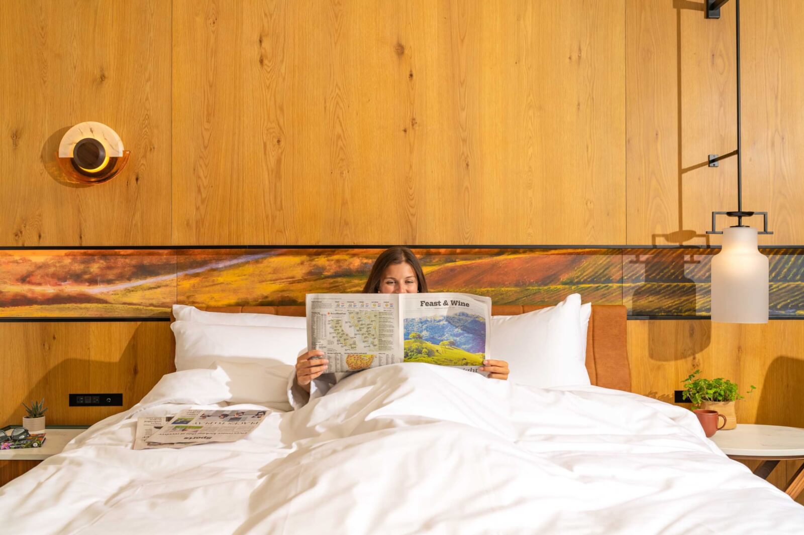 A woman relaxes in a cozy, modern bedroom with wooden walls, reading a newspaper while tucked under white sheets in Healdsburg, California.