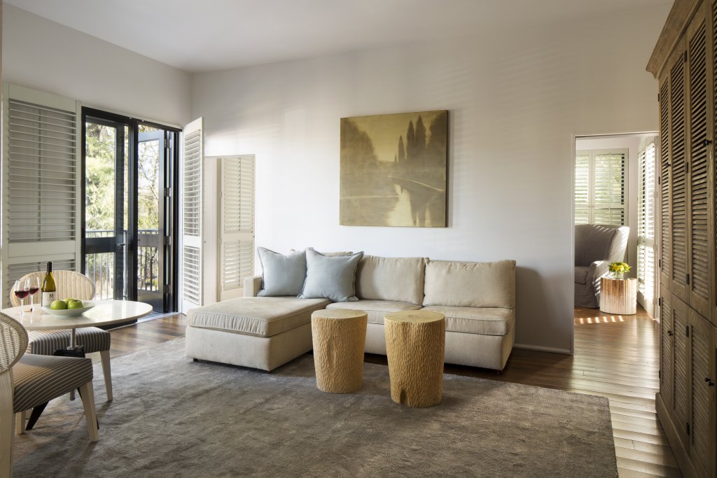 A spacious living room with a large window and sliding glass doors leading to a balcony, featuring a comfortable sectional sofa, two wooden stump side tables, a round dining table with chairs, and a bottle of wine with glasses on the table in Healdsburg, California.