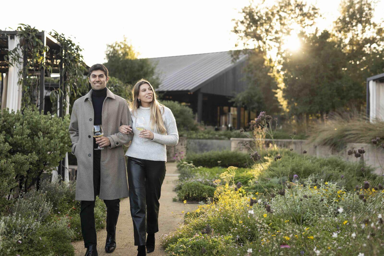 A couple strolls through a lush garden at sunset, enjoying a glass of wine together in Healdsburg, California.