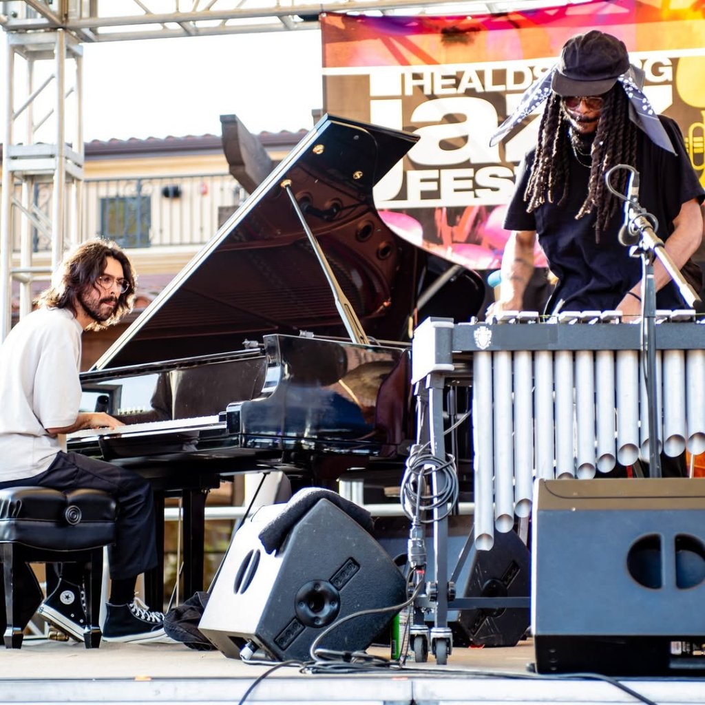 Two musicians perform at a jazz festival, one playing a grand piano and the other a keyboard and vibraphone, with a colorful banner in the background in Healdsburg, California.