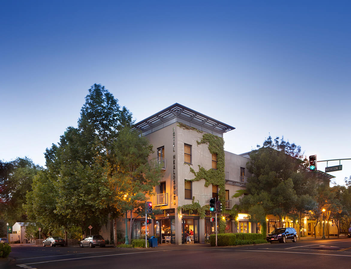 A modern building with a flat roof and large windows is nestled among lush green trees, illuminated by warm streetlights at dusk in Healdsburg, California.