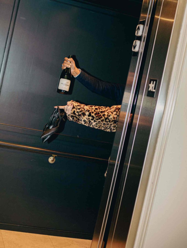 A person in a leopard print sleeve and holding a bottle of champagne emerges from an elevator, with one black high-heeled shoe dangling from their hand in Healdsburg, California.