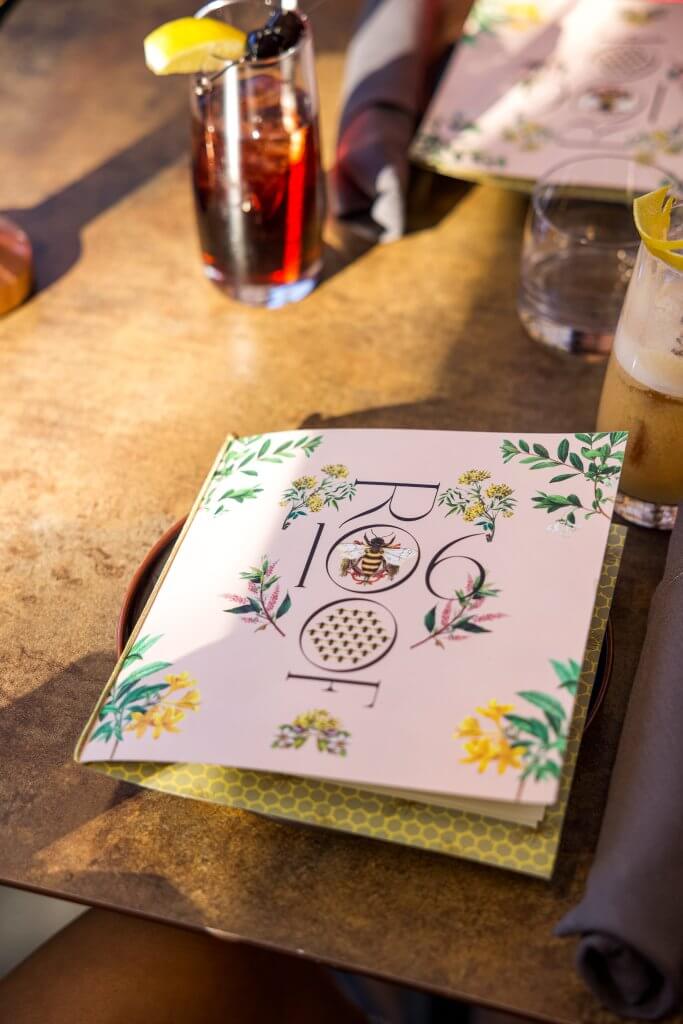 A beautifully presented menu with floral illustrations and a honey bee design sits on a wooden table alongside a glass of dark red cocktail garnished with a lemon wedge and a cherry in Healdsburg, California.