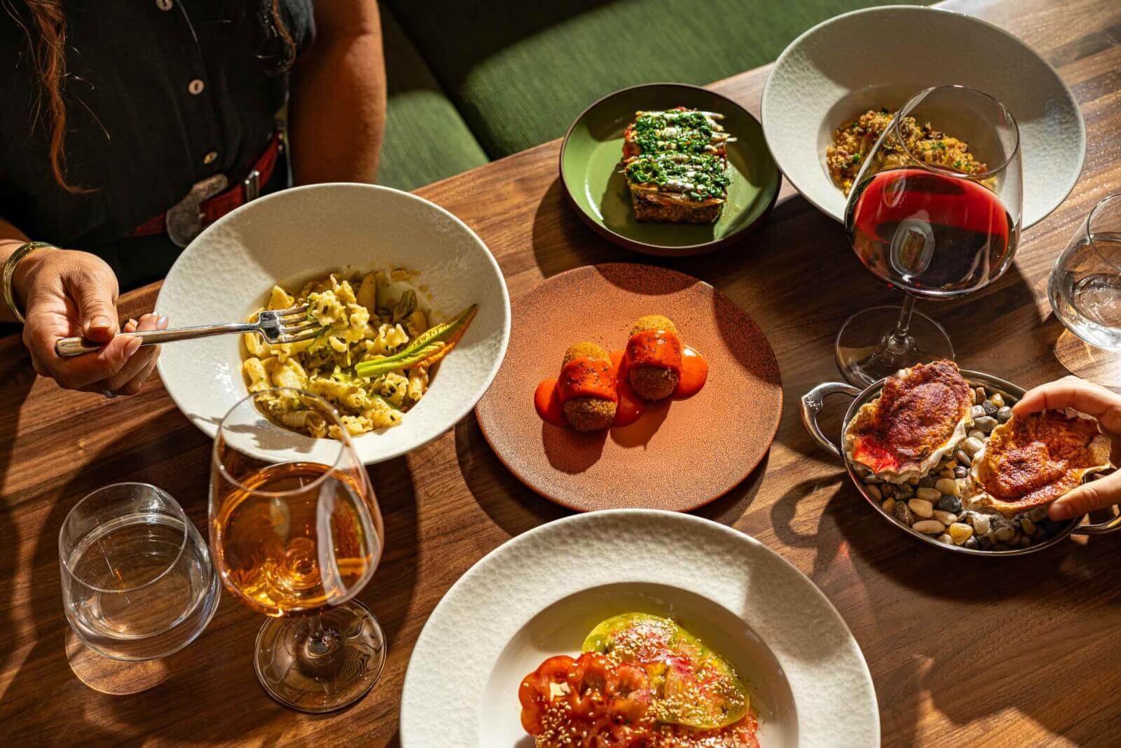 A person is enjoying a variety of Mediterranean dishes, including pasta, fried snacks, and seafood, accompanied by glasses of wine and water on a wooden table in Healdsburg, California.