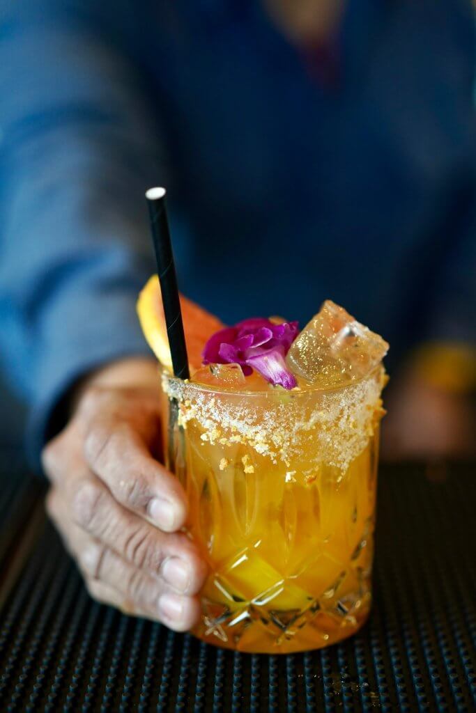A hand holds a beautifully crafted cocktail in a diamond-cut glass, garnished with a purple flower, orange slice, and a black straw in Healdsburg, California.