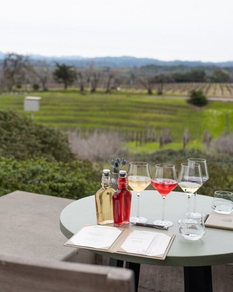 A round table set with wine glasses, bottles, and menus overlooks a picturesque vineyard and distant mountains in Healdsburg, California.