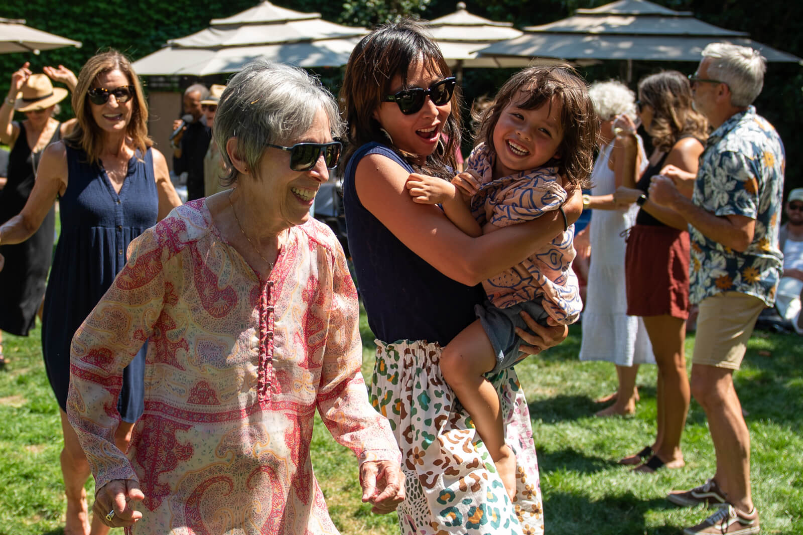 A joyful gathering of people of all ages dancing and celebrating outdoors under large umbrellas on a sunny day in Healdsburg, California.
