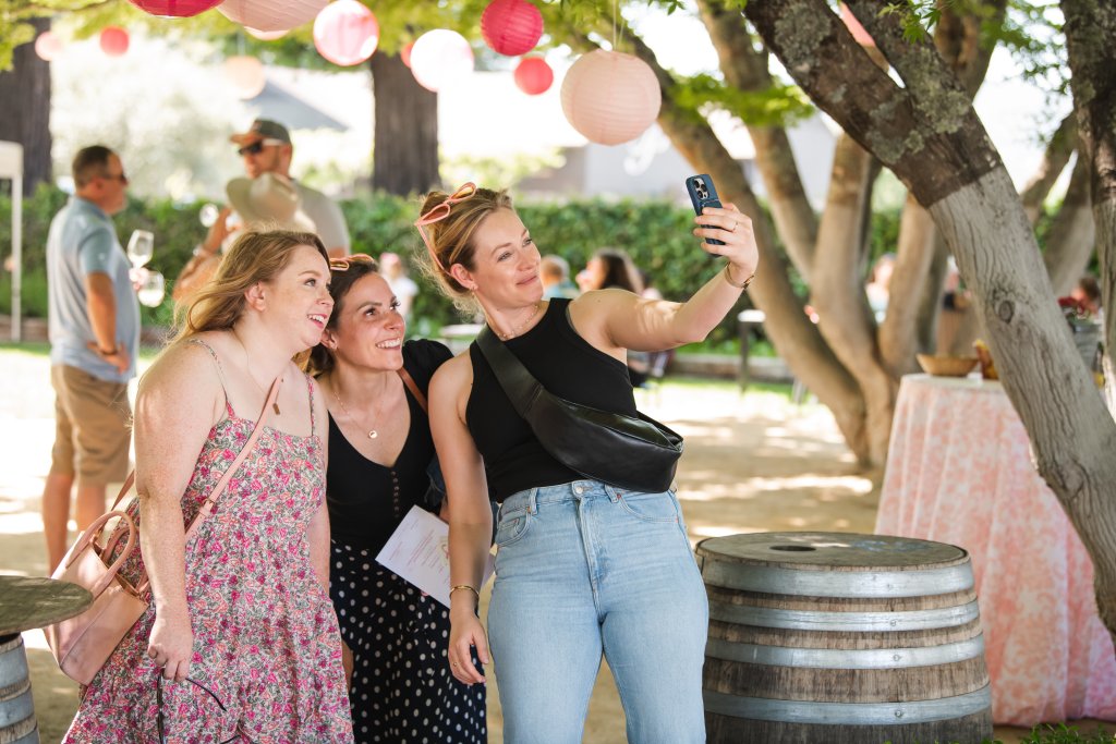 Three women are taking a selfie together at an outdoor event, surrounded by trees and hanging lanterns in Healdsburg, California.