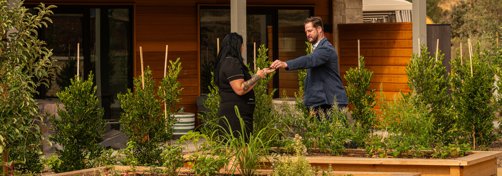 A man and woman stand in a lush garden, surrounded by wooden planters and greenery, shaking hands in front of a modern wooden house in Healdsburg, California.