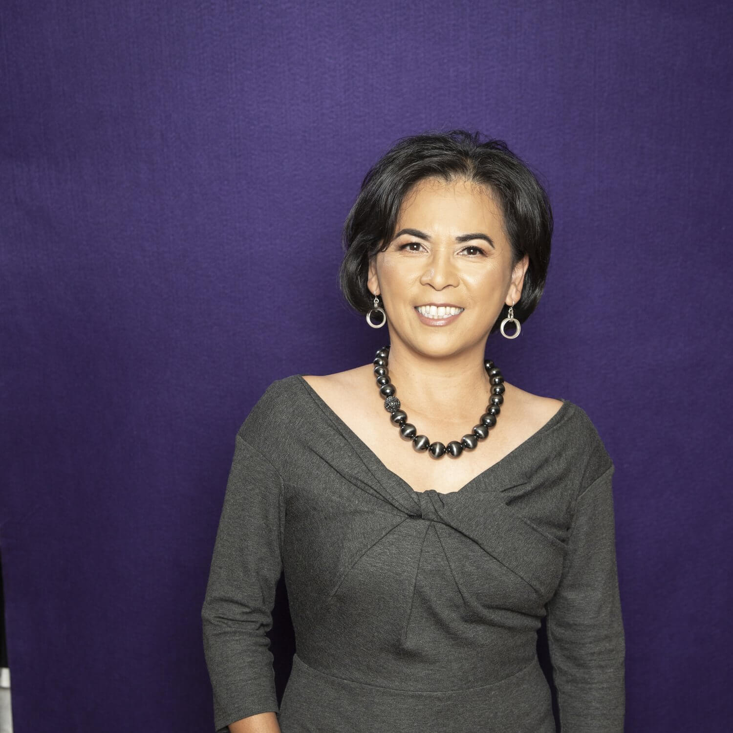 A woman with short dark hair is smiling warmly at the camera, wearing a stylish gray dress and a beaded necklace against a vibrant purple background in Healdsburg, California.