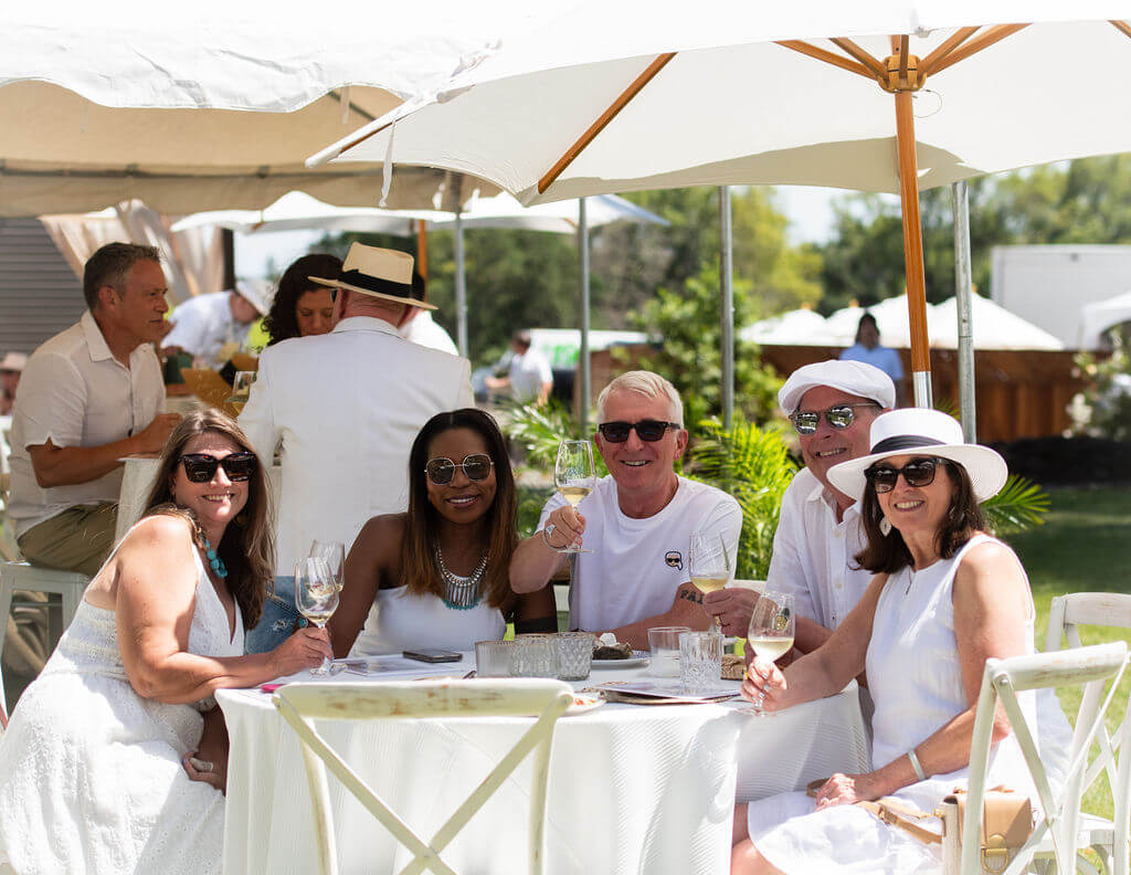 A group of elegantly dressed people enjoy a sunny outdoor brunch under large white umbrellas, raising their glasses in a toast in Healdsburg, California.