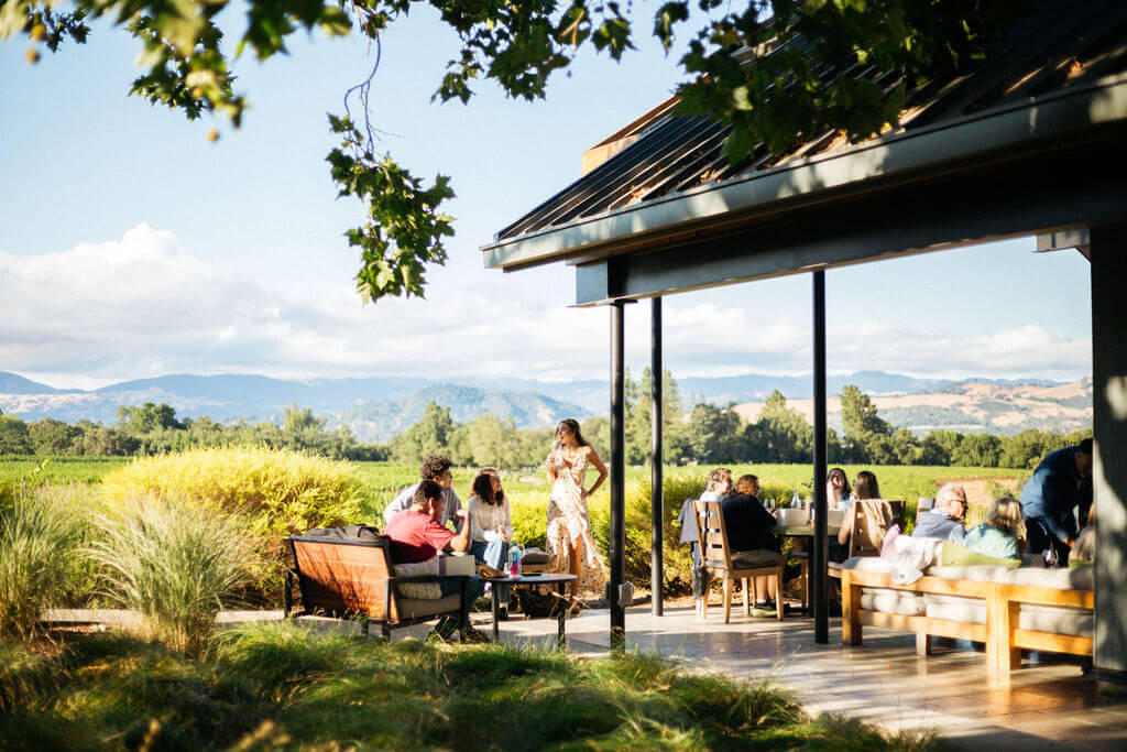 A group of people enjoys a sunny day on a patio overlooking a picturesque vineyard and distant mountains in Healdsburg, California.
