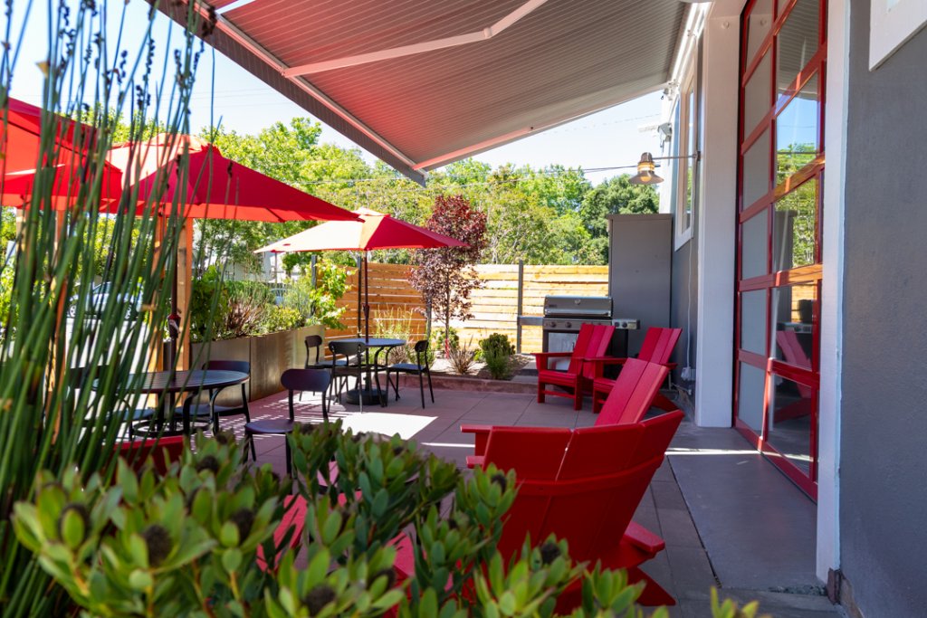 A vibrant outdoor patio area with red Adirondack chairs, tables, and umbrellas, surrounded by lush greenery and a wooden fence in Healdsburg, California.