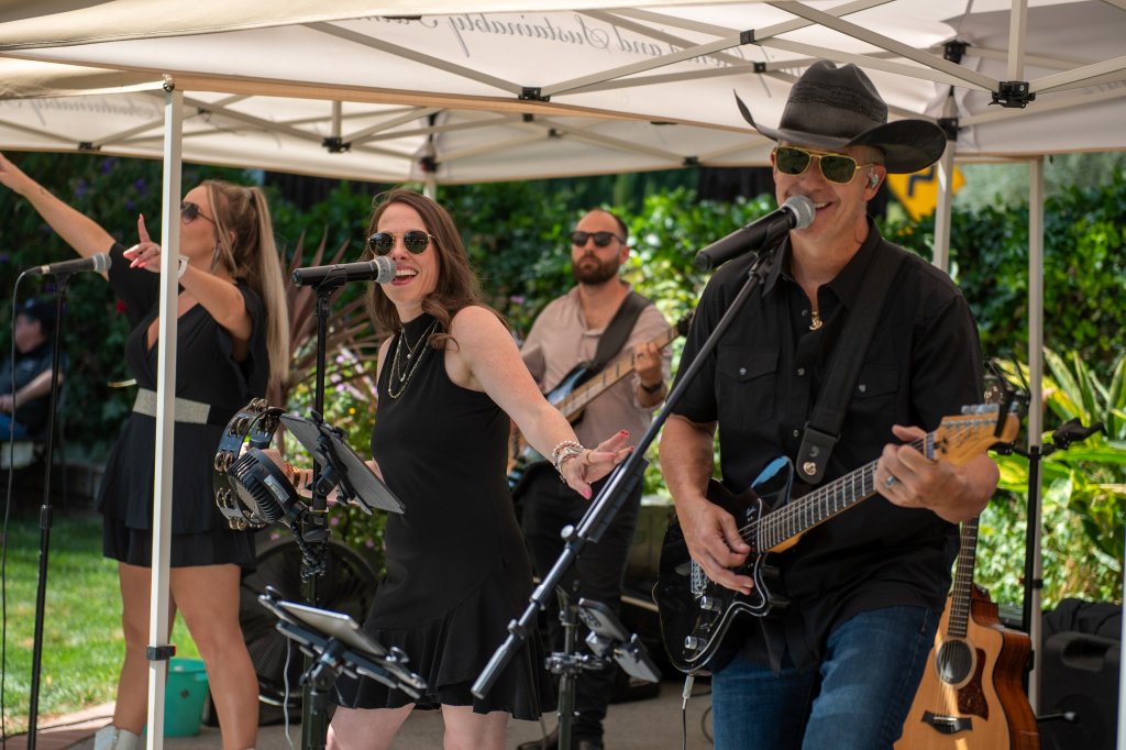 A lively outdoor concert featuring a band of five musicians performing under a white tent, with lush greenery in the background in Healdsburg, California.