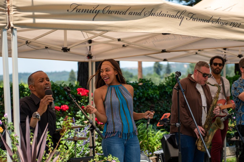 A lively outdoor concert under a white tent with musicians performing and singing, surrounded by lush greenery and colorful flowers in Healdsburg, California.