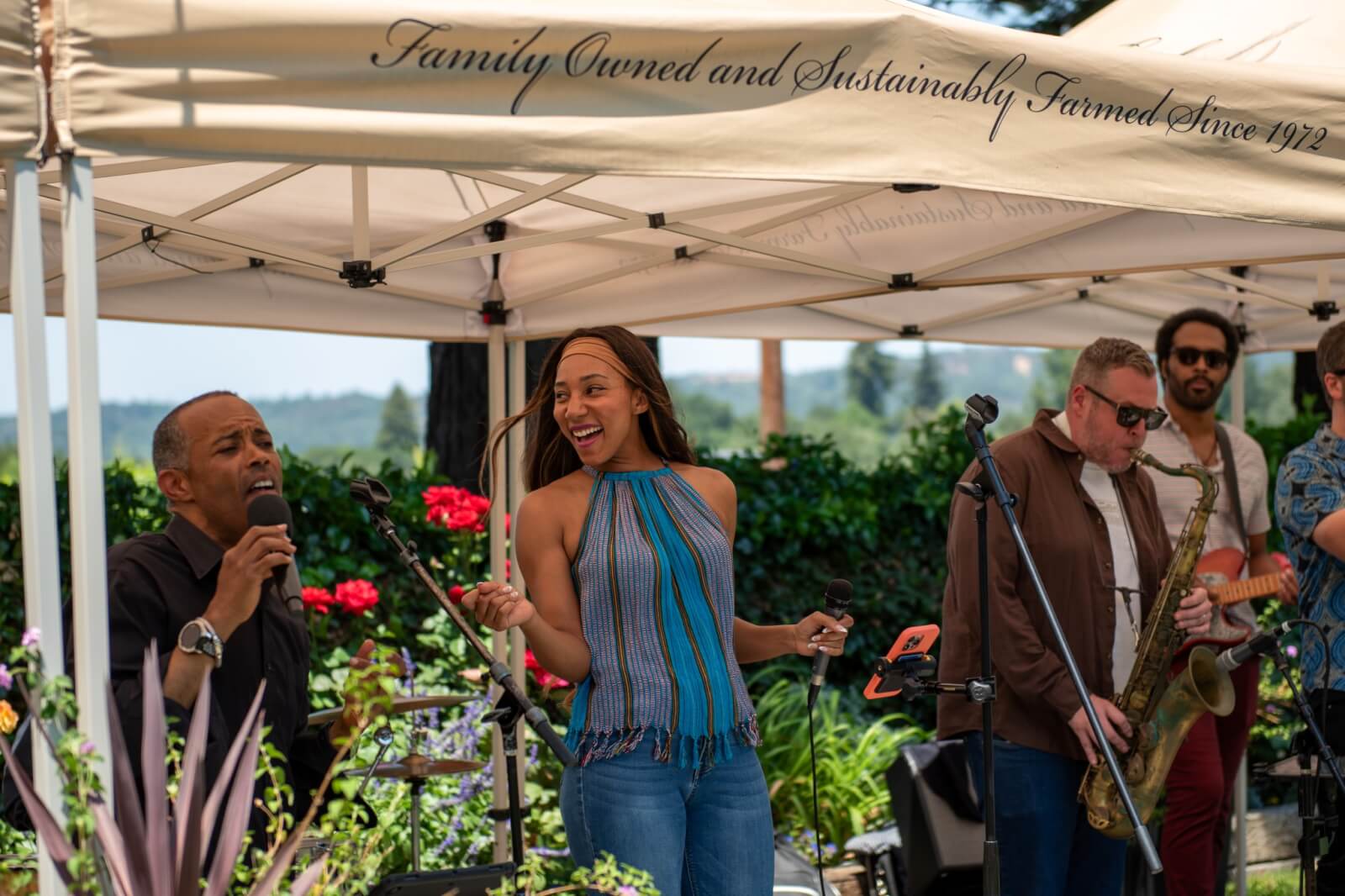 A lively outdoor concert under a white tent with musicians performing and singing, surrounded by lush greenery and colorful flowers in Healdsburg, California.