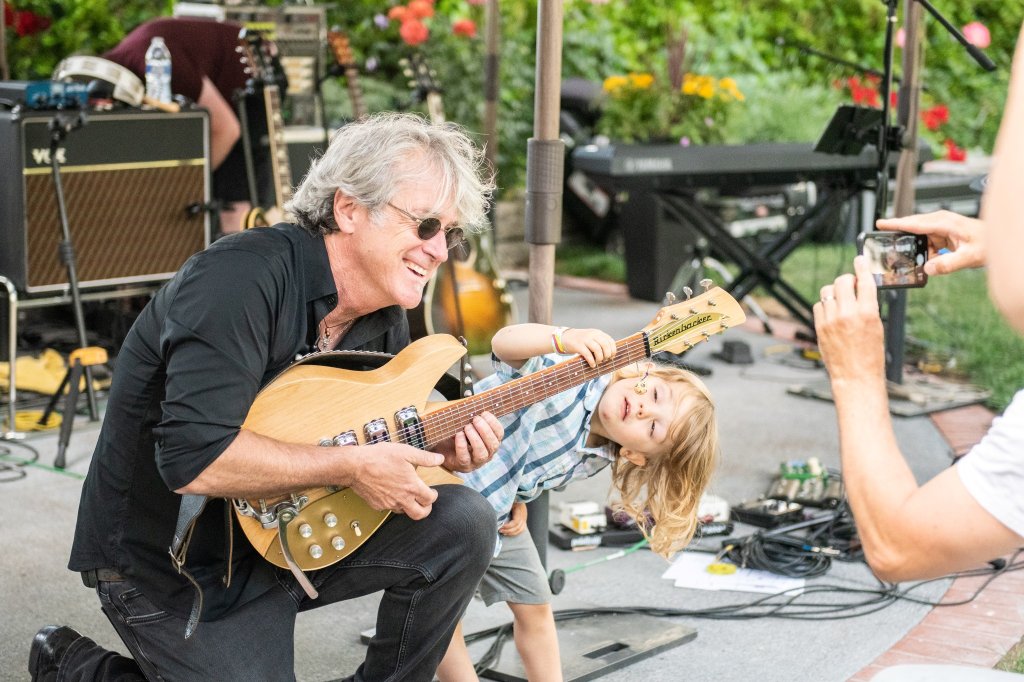 A musician with gray hair and sunglasses is joyfully playing an electric guitar with a young child, who is also holding a guitar, in a garden setting filled with musical equipment and flowers in Healdsburg, California.