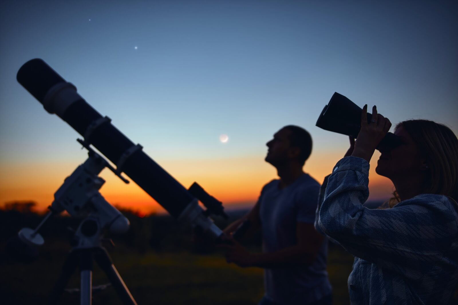 Two people are silhouetted against a vibrant sunset, observing the night sky through a telescope and binoculars, with a crescent moon and stars visible above in Healdsburg, California.