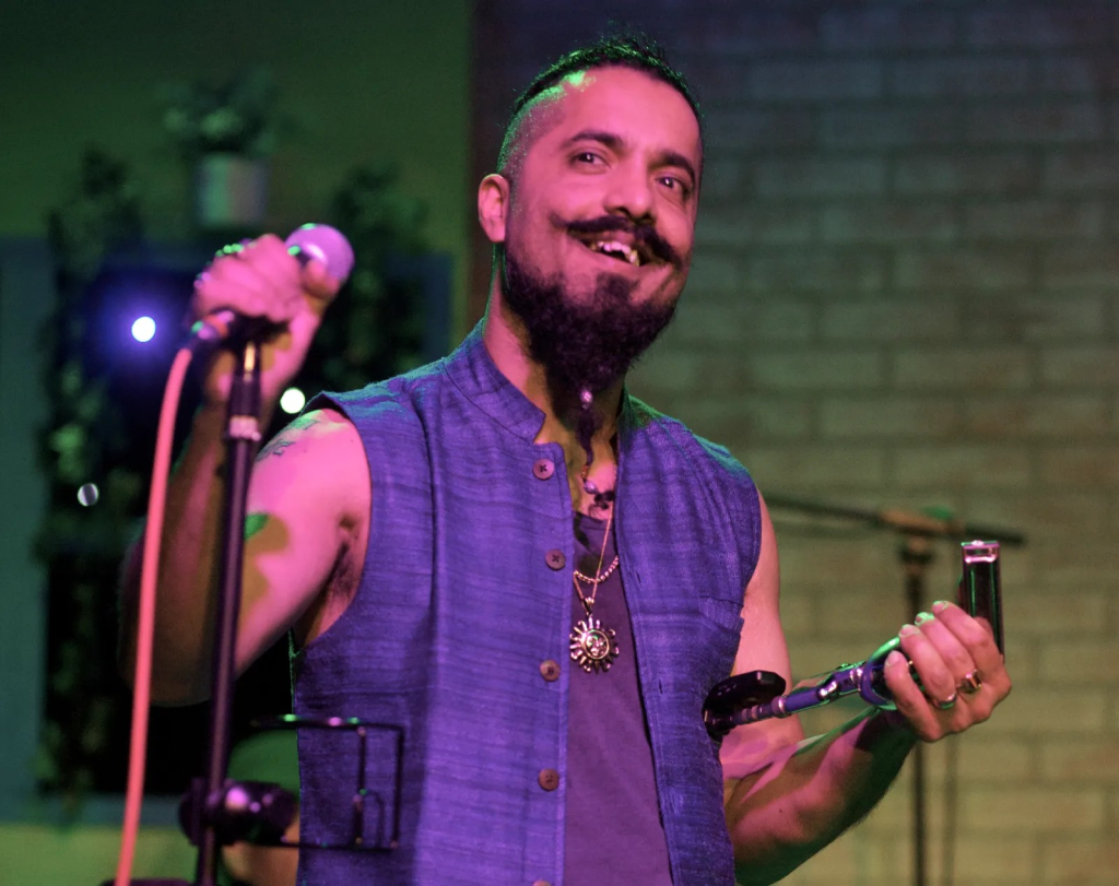 A man with a beard and mustache, wearing a purple sleeveless shirt, performs on stage with a microphone in one hand and a harmonica in the other in Healdsburg, California.