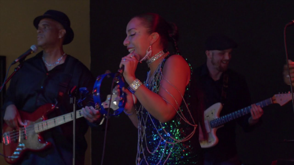 A vibrant musical performance featuring a female singer in a sparkling dress, a guitarist, and a bassist, all smiling and engaged in their music in Healdsburg, California.