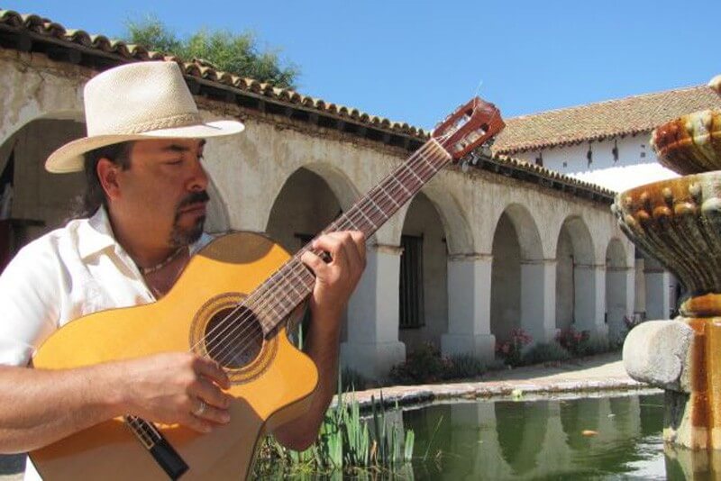 A musician wearing a straw hat plays an acoustic guitar in a serene courtyard with an ornate fountain and arched walkways in Healdsburg, California.