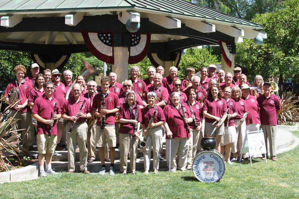 A large group of musicians in maroon shirts and khaki pants perform together under a gazebo decorated with American flags in Healdsburg, California.