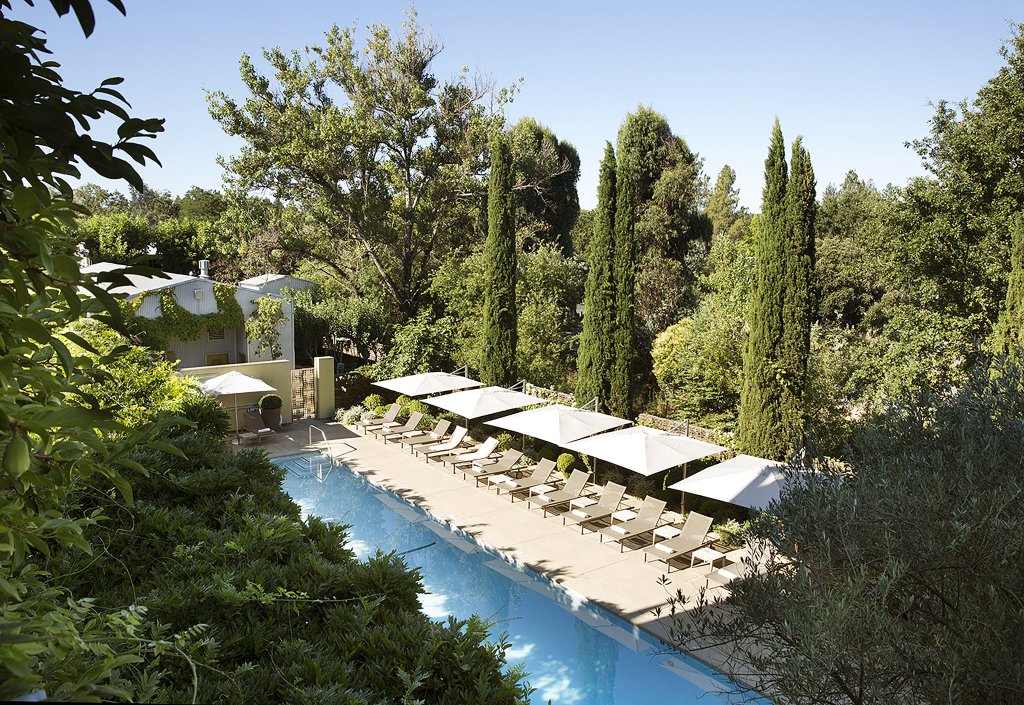 A serene outdoor swimming pool surrounded by lush greenery and shaded by white umbrellas, with a nearby building partially obscured by trees in Healdsburg, California.