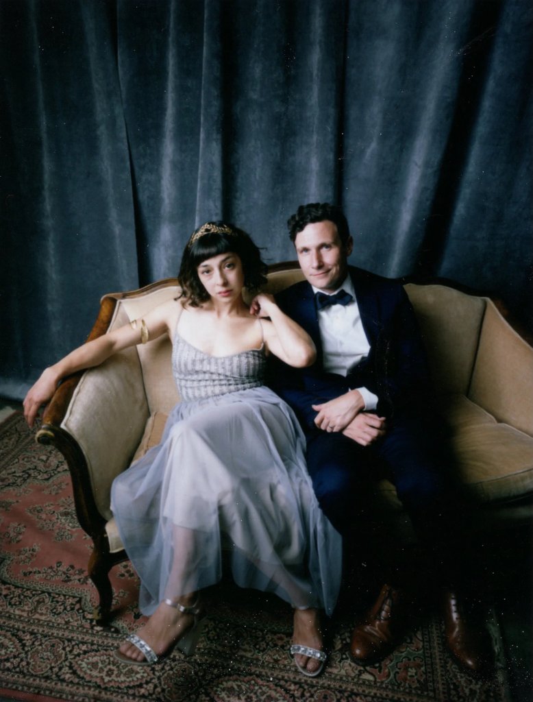 A couple dressed in formal attire sits elegantly on a vintage sofa against a dark curtain backdrop in Healdsburg, California.