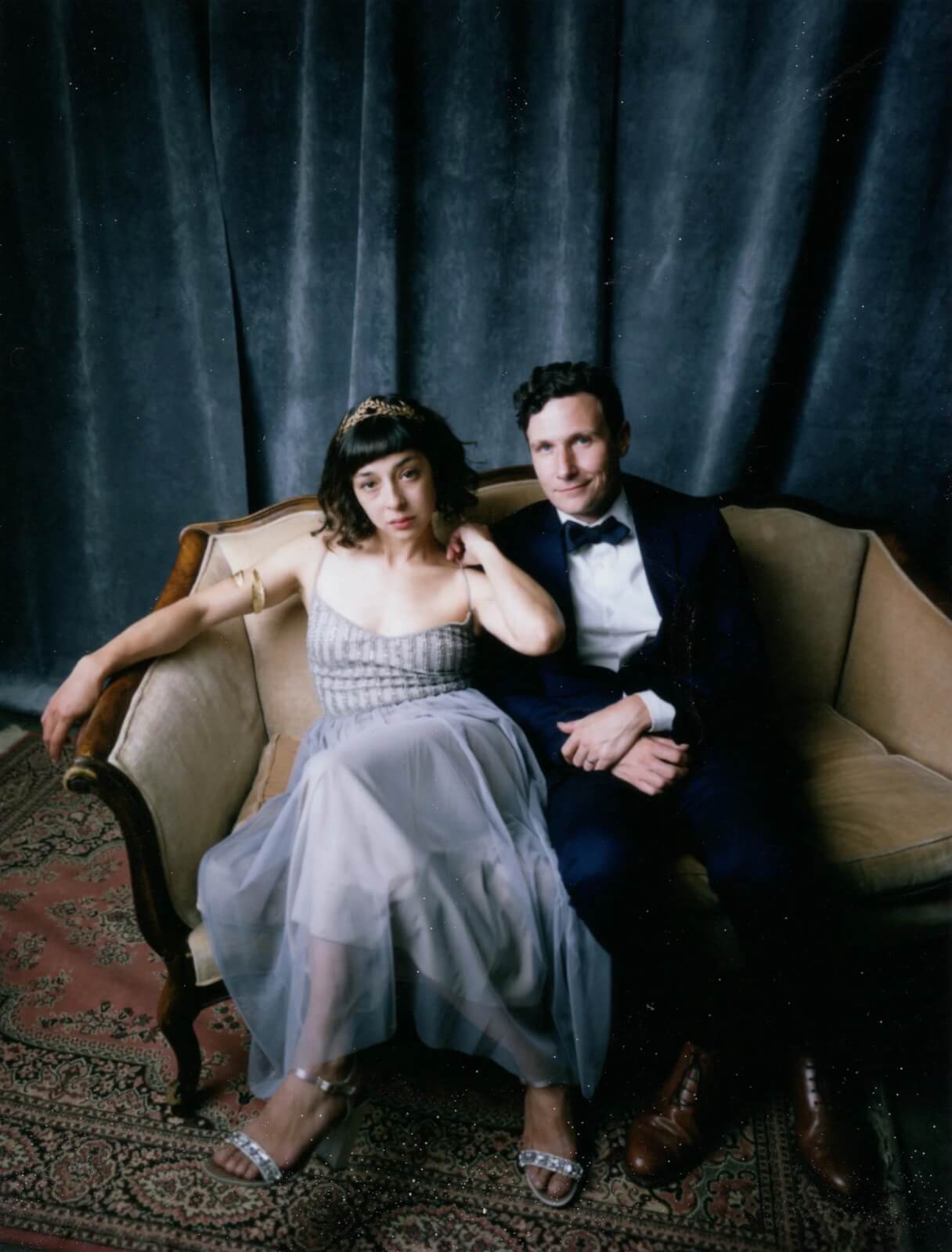 A couple dressed in formal attire sits elegantly on a vintage sofa against a dark curtain backdrop in Healdsburg, California.