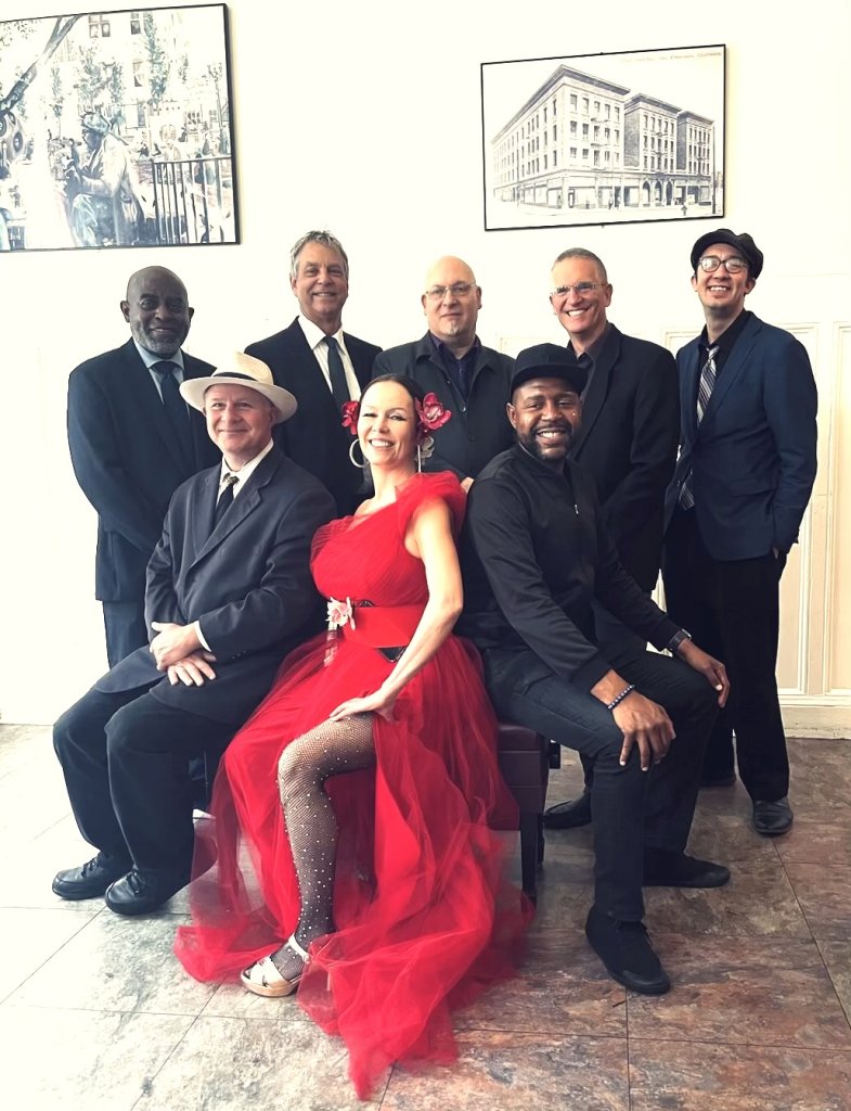 A group of eight elegantly dressed individuals, including a woman in a striking red gown, pose together in a formal setting with framed artwork on the walls in Healdsburg, California.