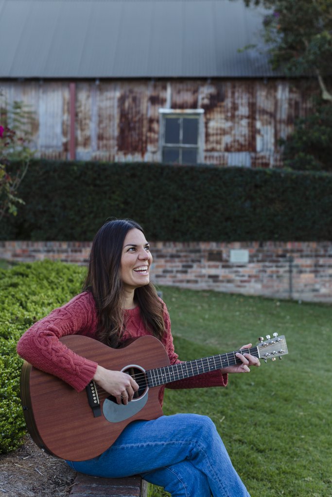 A woman with long dark hair sits on a bench, smiling as she plays an acoustic guitar in a garden setting in Healdsburg, California.