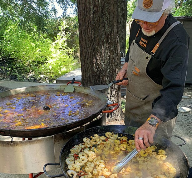 A chef in an apron and hat is cooking a large paella with seafood and vegetables outdoors in Healdsburg, California.