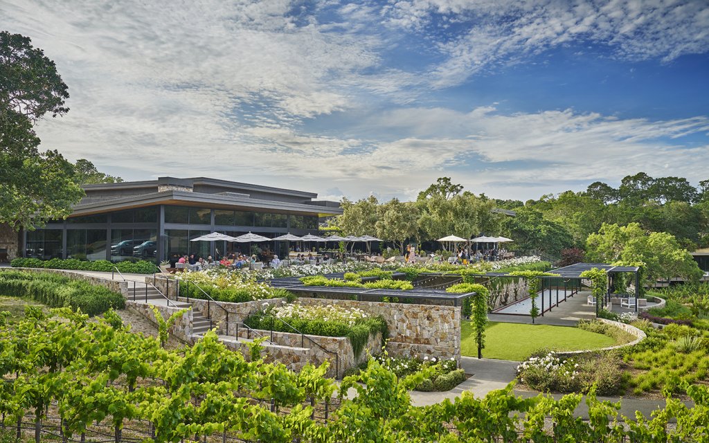 A sprawling outdoor dining area with stone walls and lush gardens overlooks a picturesque vineyard and modern building in Healdsburg, California.