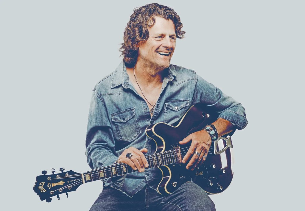 A musician with curly hair, wearing a denim shirt and jeans, smiles while playing an electric guitar in Healdsburg, California.