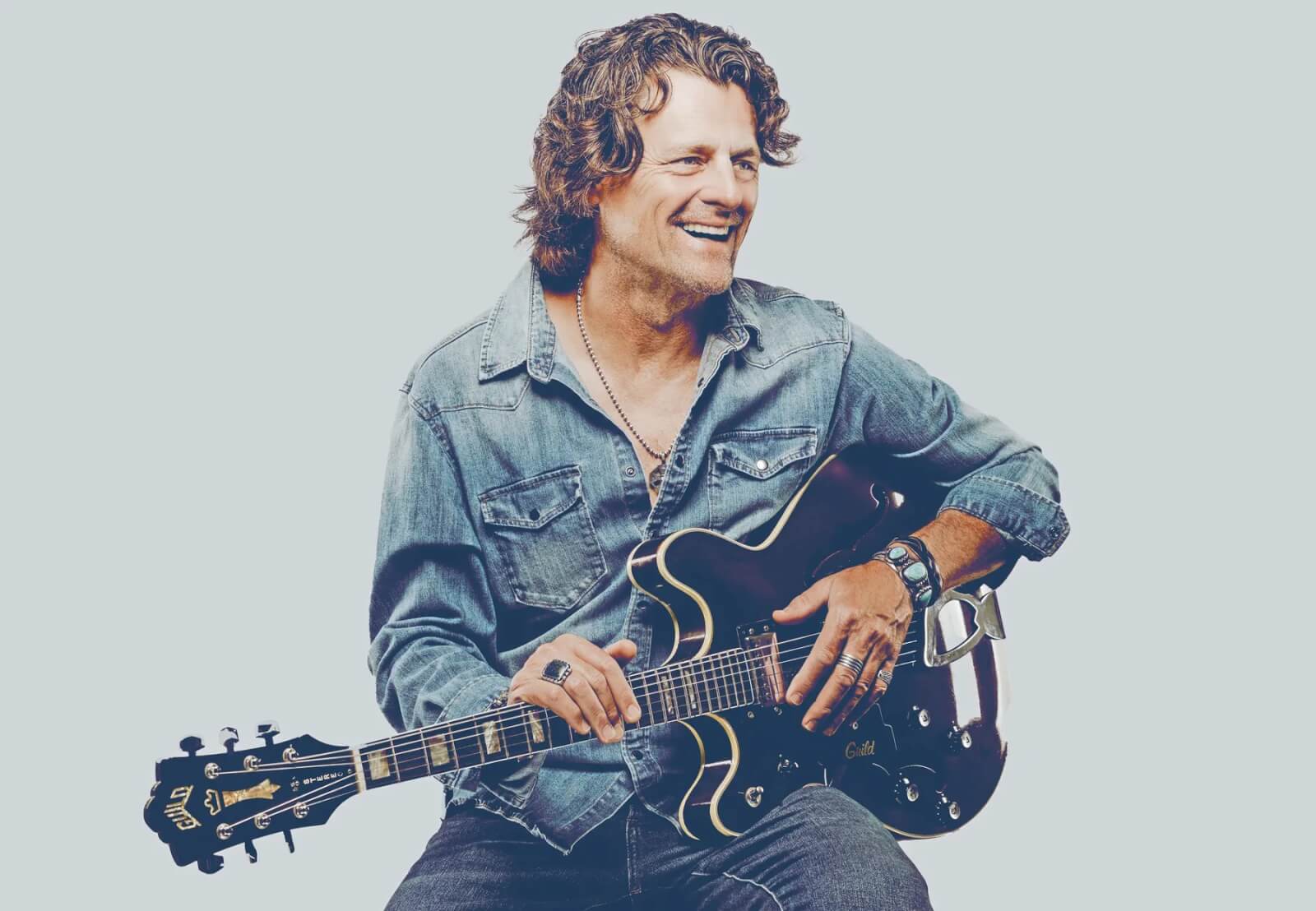A musician with curly hair, wearing a denim shirt and jeans, smiles while playing an electric guitar in Healdsburg, California.