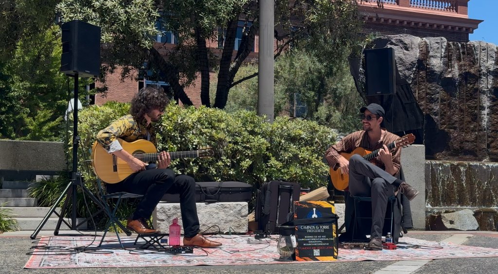 Two musicians play acoustic guitars on a colorful rug in an outdoor setting with lush greenery and a brick building in the background in Healdsburg, California.