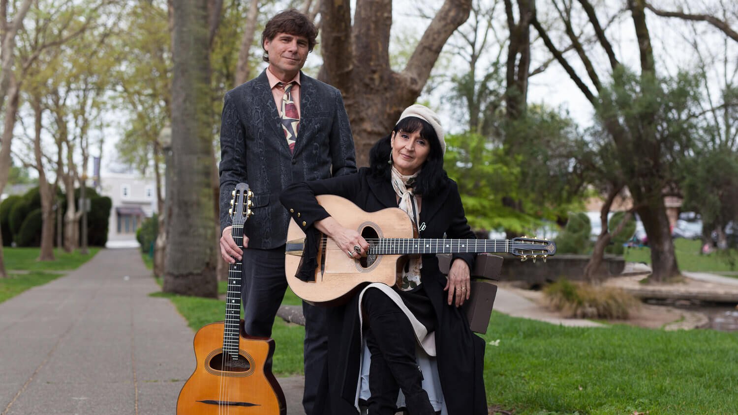 A man and woman dressed in stylish black attire pose with their guitars in a serene park setting in Healdsburg, California.