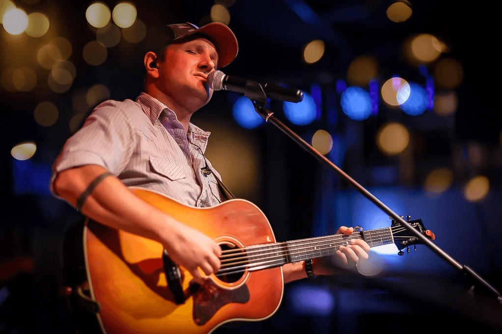 A musician passionately sings into a microphone while playing an acoustic guitar on stage, illuminated by warm stage lights in Healdsburg, California.