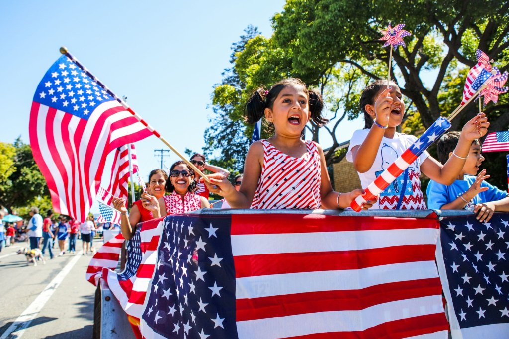 A vibrant Fourth of July parade float adorned with American flags, featuring enthusiastic children waving flags and holding patriotic pinwheels, with a backdrop of cheering spectators and a clear blue sky in Healdsburg, California.