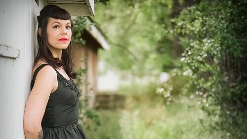 A woman in a black dress stands against a weathered white shed, surrounded by lush greenery in Healdsburg, California.