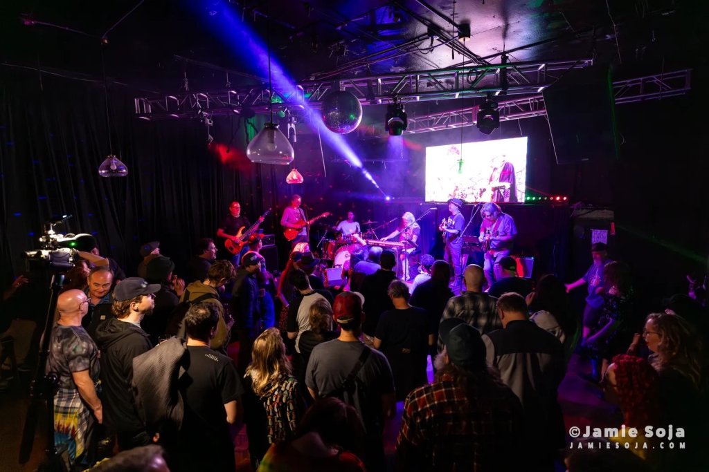 A lively concert scene with a band performing on stage, illuminated by colorful stage lights, while an enthusiastic crowd watches and dances in the dimly lit venue in Healdsburg, California.