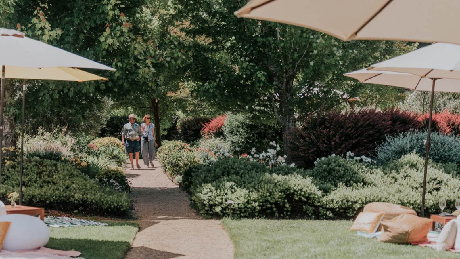 Plan Your Trip A couple strolls down a garden path lined with lush greenery and colorful flowers, shaded by large umbrellas in Healdsburg, California.