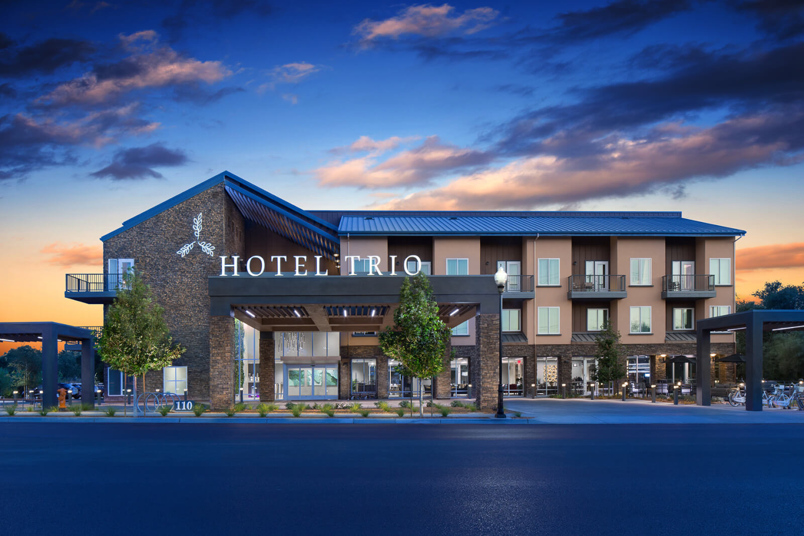 Hotel Trio healdsburg A modern hotel building named Hotel Trio with a distinctive architectural design featuring a slanted roof and large windows, set against a backdrop of a colorful sunset sky in Healdsburg, California.